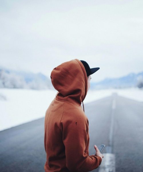 A person in a hoody stands on a snowy road, holding a smartphone, with mountains in the background.